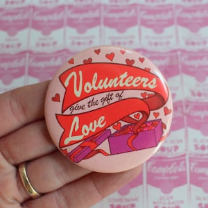May include: A pink button with the text "Volunteers give the gift of Love" in red and white. The button has heart and gift box illustrations. The background is a pink pattern of soup cans.