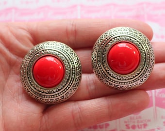 May include: A pair of silver tone clip-on earrings with a red cabochon stone in the center of each earring. The earrings have a decorative, ornate design.
