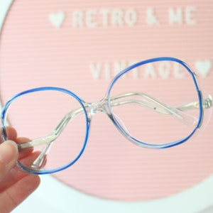 May include: Clear blue-rimmed glasses with a silver frame. The glasses are held in a hand against a pink background with the text "Retro & Me Vintage" on it.