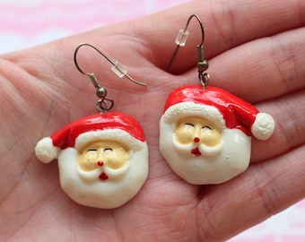 May include: A pair of silver dangle earrings with a white and red Santa Claus face charm. The Santa Claus face is smiling with closed eyes.