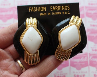 May include: A pair of black and white square earrings with gold accents. The earrings feature a white diamond-shaped stone in the center. The earrings are labeled "Fashion Earrings Made in Taiwan R.O.C."
