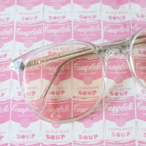 Puede incluir: Gafas con montura de plástico transparente y patillas de metal dorado, descansando sobre un fondo rosa y blanco con imágenes repetidas de latas de sopa Campbell's. Las latas presentan varios sabores de sopa, incluyendo tomate, champiñón, consommé, frijol negro y pepperpot.