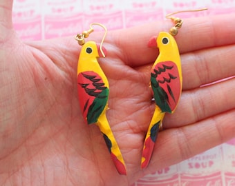 May include: A pair of wooden parrot earrings with yellow, red, and green paint. The earrings have gold hooks.