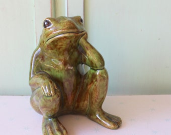 May include: A ceramic frog figurine in shades of green and brown, posed with a thoughtful expression. The frog is seated, with one hand resting on its chin. The figurine is on a pink surface with a light green background.