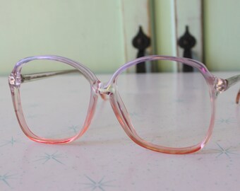 May include: Clear-framed eyeglasses with a gradient pink and orange tint on the lower rims. The glasses have a large, square shape and silver-toned arms. The lenses are clear, and the frames are made of a transparent plastic material.