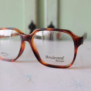 May include: Tortoise shell eyeglasses with a square frame and clear lenses. The glasses have the text "Boulevard Boutique" printed on the right lens.