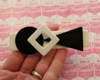 Puede incluir: Una pinza para el pelo geométrica en blanco y negro con forma cuadrada y circular. La pinza está hecha de plástico y tiene un acabado brillante.