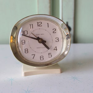 Peut inclure: Un réveil vintage Westclox blanc et or avec un cadran rond et des aiguilles noires. Le cadran de l'horloge a des chiffres noirs et le texte "Westclox" et "Sixty Six" imprimé dessus.