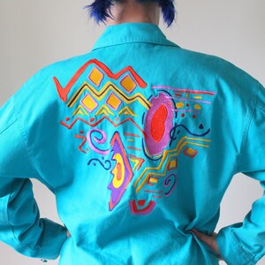 May include: A turquoise denim jacket with a colorful abstract design embroidered on the back. The design features geometric shapes, swirls, and lines in shades of red, yellow, orange, pink, and purple.