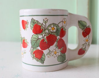May include: White ceramic mug with a handle, featuring a strawberry pattern. The mug has a green border and red strawberries with green leaves and white flowers.