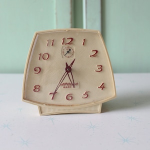 Peut inclure: Un réveil vintage blanc avec des chiffres rouges et un cadran de couleur crème. L'horloge a une petite aiguille des secondes et les mots "APOLLO MARK" imprimés sur le cadran.