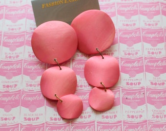 May include: A pair of pink statement earrings with a layered design. The earrings are made of metal and have a smooth, matte finish.