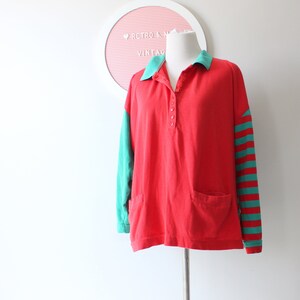 May include: Red long-sleeved shirt with a green and white striped sleeve and a green collar. The shirt has a button-up front and a pocket on the left side.