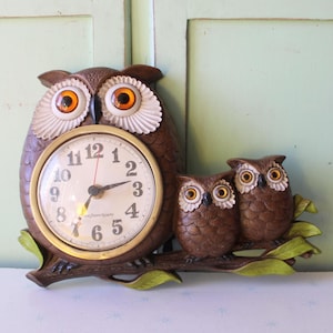 May include: A brown owl-shaped clock with a gold-colored clock face and two smaller owl figurines. The clock face has black numbers and hands. The owls are perched on a brown branch with green leaves. The clock measures approximately 12 inches in height.