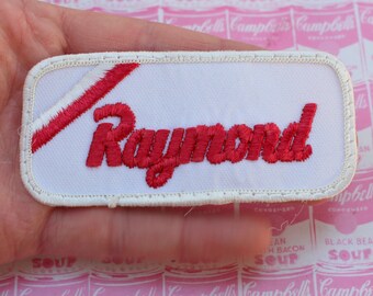 May include: White embroidered patch with a red diagonal stripe and the word "Raymond" in red cursive lettering.