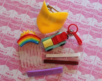 Könnte beinhalten: Eine Sammlung bunter Haarschmuckstücke, darunter ein Regenbogenkamm, eine gelbe Haarklammer, ein rotes und blaues Spielzeugauto und ein lila Haargummi. Die Accessoires sind auf einem rosa und weißen Hintergrund mit einem Muster von Campbell's Suppendosen angeordnet.