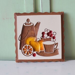 May include: A brown wooden framed tile with a white background featuring a brown teapot, two brown cups, a yellow lemon, and a bunch of red grapes.
