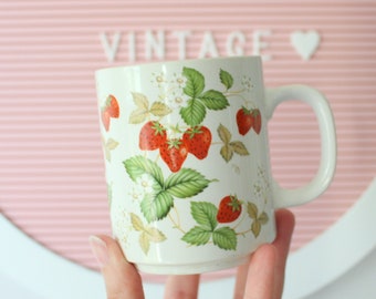 May include: White ceramic mug with a strawberry pattern. The mug has a handle and is decorated with red strawberries, green leaves, and white flowers.