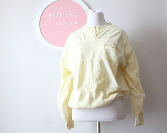 May include: A pale yellow long-sleeved shirt with a button-up collar and a single pocket on the left side. The shirt is on a white mannequin against a white wall.