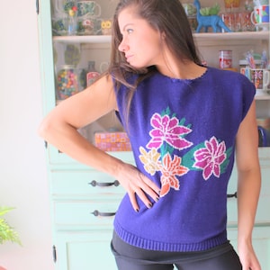Op de afbeelding: Een vrouw draagt een paarse, mouwloze trui met een bloemenpatroon. De trui heeft een ronde hals en is gemaakt van een zachte, gebreide stof. De bloemen zijn geborduurd in tinten roze, geel en groen.