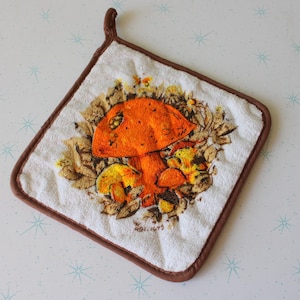 Puede incluir: Un agarrador de olla marrón y blanco con un diseño de hongo. El hongo es naranja con manchas marrones y está rodeado de hojas verdes. El agarrador de olla tiene un borde marrón.