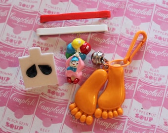 May include: A set of colorful plastic charms featuring a pair of orange flip flops, a clown, and a bunch of balloons. The charms are on a pink background with a pattern of Campbell's Soup cans.