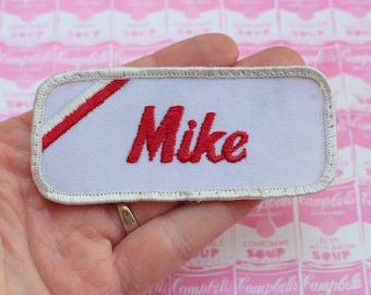 May include: White embroidered iron-on patch with a red diagonal stripe and the name "Mike" in red lettering.