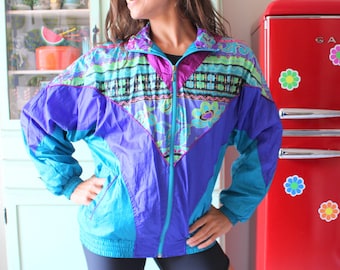 Può includere: Una giacca a vento con motivo blu, viola e verde brillante con chiusura a zip. La giacca ha un design geometrico con un motivo floreale.