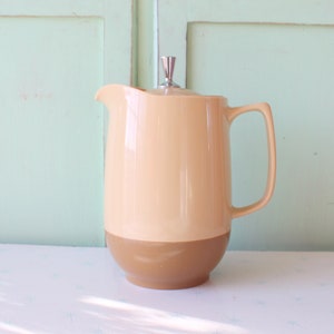 Puede incluir: Una jarra de cerámica beige y marrón con tapa y asa plateadas. La jarra está sobre una superficie blanca.