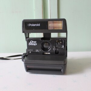 Peut inclure: Un appareil photo instantané Polaroid One Step noir avec un flash intégré. L'appareil photo est doté d'une sangle noire.