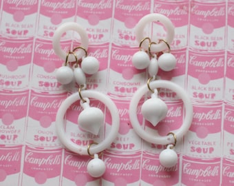 May include: A pair of white plastic earrings with multiple circles and white beads. The earrings are hanging against a pink background with a repeating pattern of Campbell's Soup cans.
