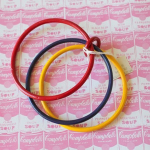 May include: Four interlocking wooden rings in red, yellow, and blue, arranged in a circle on a pink and white background with a repeating pattern of Campbell's Soup cans.