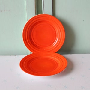 May include: Two orange ceramic plates with a concentric circle design. The plates are stacked on top of each other on a white surface with a light blue pattern.