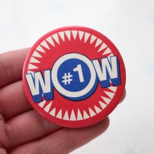 May include: A round, red button with the word "WOW" in white and blue lettering. The number "1" is inside a blue circle. The button has a white, jagged border. The button is held in a hand.