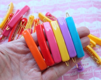 May include: A set of colorful plastic barrettes in orange, red, pink, yellow, purple, and blue. The barrettes are arranged in a hand, with the gold metal clips facing up.