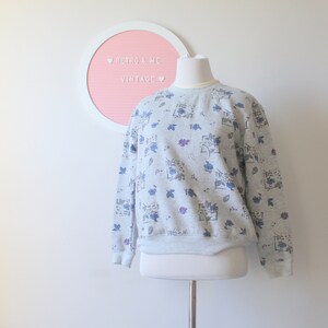May include: A light gray vintage sweatshirt with a floral pattern in blue and purple. The crew neck sweatshirt is displayed on a mannequin. A pink letter board with white text is in the background.