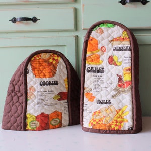 Puede incluir: Dos fundas acolchadas marrones para electrodomésticos de cocina. Una es arqueada y la otra rectangular. Ambas tienen un centro blanco con ilustraciones coloridas de productos horneados y recetas. La funda arqueada dice "COOKIES" y la rectangular dice "CAKES" y "ROLLS".