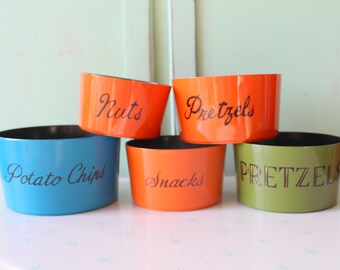 May include: Five colorful snack bowls in blue, orange, and green, each with a black interior. The bowls are labeled with the words "Potato Chips", "Snacks", "Nuts", "Pretzels", and "Pretzels" in black script.