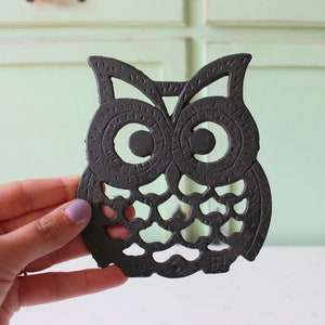 Puede incluir: Posavasos de hierro fundido negro en forma de búho. El búho tiene ojos grandes y redondos y detalles intrincados.
