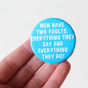 May include: A round, light blue button with white text that reads: "MEN HAVE TWO FAULTS, EVERYTHING THEY SAY AND EVERYTHING THEY DO!" The button is held by a hand.