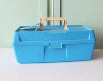 Puede incluir: Una caja de herramientas de plástico turquesa vintage con asa y cierre beige. La caja rectangular tiene una superficie texturizada y está sobre una mesa blanca. El fondo presenta una puerta verde claro.