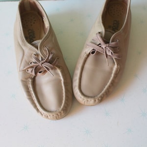 May include: A pair of beige leather casual shoes with laces. The shoes have a moccasin-style toe and are tied with brown laces.