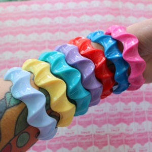 May include: A set of colorful, wavy plastic bracelets in shades of blue, yellow, purple, red, and pink. The bracelets are stacked on a person's arm.
