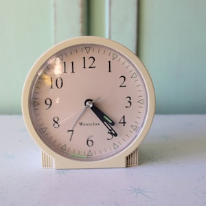 Puede incluir: Un reloj despertador analógico blanco con una esfera rosa y agujas negras. La esfera del reloj tiene números negros y la palabra "Westclox" impresa en ella.