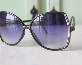 Puede incluir: Gafas de sol negras con un borde dorado alrededor de las lentes. Las gafas tienen una montura grande y redondeada.