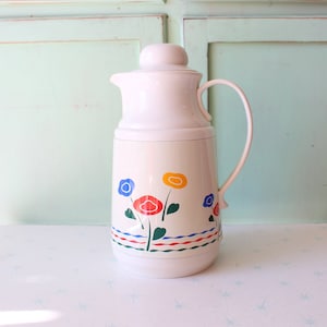 May include: White thermal carafe with a floral design featuring red, blue, yellow, and green flowers. The carafe has a lid and a handle.