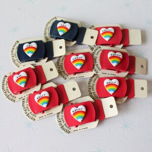Könnte beinhalten: Eine Sammlung roter und blauer, herzförmiger Gürtelschnallen aus Kunststoff mit Regenbogenakzenten. Jede Schnalle trägt einen anderen Namen, darunter Karen, Kathy, Kelly und Kim. Die Schnallen sind auf weißen Karten mit dem Text "Ceramic Figure Buckle" und "Heart Pin Buckle" verpackt.