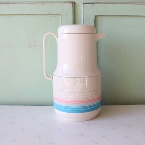 May include: White thermal carafe with a blue, pink and white striped design. The carafe has a lid and a handle.