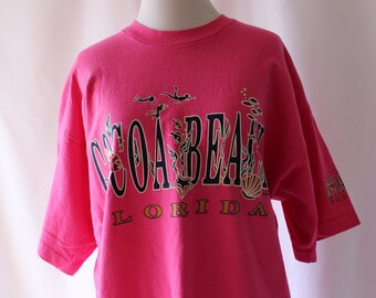 May include: Pink short-sleeved t-shirt with a graphic print of "COCOA BEACH FLORIDA" in black and gold lettering with a seashell and seahorse design.