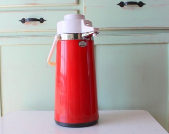 May include: A vintage red and white thermos with a handle. The thermos has a white top and a red body. It is sitting on a white table in front of a light green cabinet.